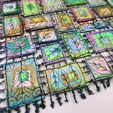 Detail of piece made up of colorful squares connected with thread decorated with embroidered leaf designs.
