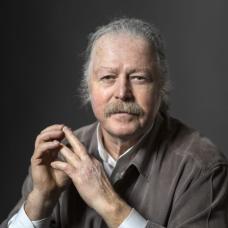 Portrait of a man with his hair tied back and a mustache in a button up shirt sits at a table